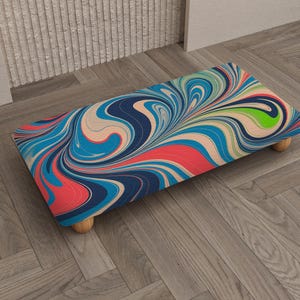 May include: A rectangular footstool with a vibrant, swirled pattern in shades of blue, red, and green. The design resembles flowing paint or liquid. The stool has four small, rounded wooden legs.