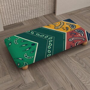 May include: A rectangular footstool with a colorful paisley pattern in green, yellow, red, and blue. The footstool has four wooden legs and is placed on a wooden floor.