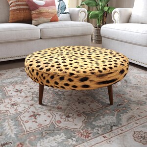May include: A round ottoman featuring a tan and black leopard print pattern across its top and sides. The piece is supported by four tapered wooden legs in a medium brown finish, providing a stable base for this circular furniture item.
