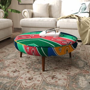 Puede incluir: Un otomán redondo con un estampado de bandana paisley multicolor en tonos rojo, verde, azul y amarillo. La pieza se apoya sobre cuatro patas de madera cónicas e incluye un borde ribeteado en amarillo a lo largo de la parte superior.