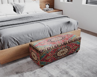 Puede incluir: Un banco de almacenamiento rectangular con una parte superior estampada colorida, roja, dorada y turquesa, se encuentra al pie de una cama. El banco tiene patas de madera. La cama tiene un edredón y almohadas grises claros.
