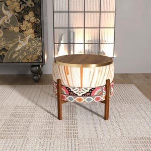 May include: A round, decorative ottoman with a wooden top and base. The top is a light brown color with a gold-colored trim. The sides are patterned with orange and white stripes, and a colorful floral design. The ottoman is supported by a dark brown wooden frame.