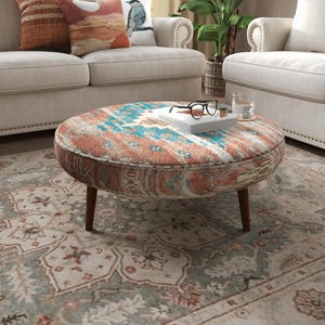 May include: A round ottoman featuring a patterned fabric in shades of terracotta, teal, and cream, supported by four tapered dark wood legs. A white book, a pair of black-rimmed glasses, and a clear glass of water sit on the top surface.