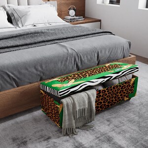 May include: A decorative storage chest with a leopard print design. The chest has a green and white zebra print lid and is open, revealing folded blankets and a gray scarf. The chest is placed at the foot of a bed.