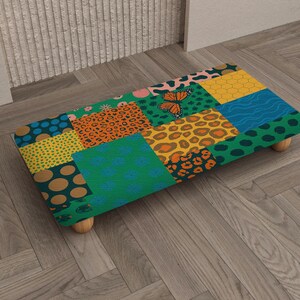 Può includere: Un pouf rettangolare con un design patchwork. Il design include stampa leopardata, pois e motivi floreali. Una farfalla è visibile sulla parte superiore. Il pouf è sostenuto da quattro piccole gambe rotonde in legno. Misura circa 60 cm di lunghezza.