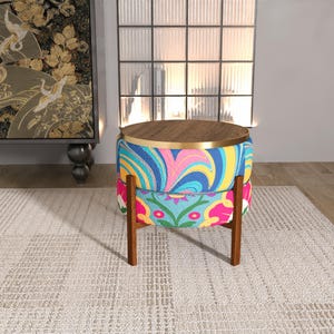 May include: A round ottoman with a wooden top and base. The ottoman's sides feature vibrant, colorful fabric with a swirling pattern of blue, pink, and yellow. The base is supported by four wooden legs.