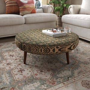 May include: A round ottoman featuring a dark green and beige geometric patterned fabric on the top surface and a tan and black abstract print on the sides. The piece is supported by four tapered wooden legs and holds a book and glasses.
