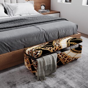 May include: A storage bench with a black, gold, and leopard print design, placed at the foot of a bed. The bench is open, revealing a gray blanket and a folded scarf. The bed has a gray comforter and white pillows.