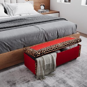 May include: A red storage bench with a leopard print top and a gray blanket draped over the side. The bench is positioned at the foot of a bed with gray bedding and white pillows. A gray rug covers the floor.