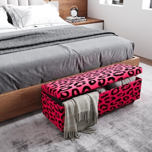 May include: A pink and black leopard print storage bench is placed at the foot of a bed with gray bedding. The bench is open, revealing folded blankets and a gray throw. The room has a neutral color palette.