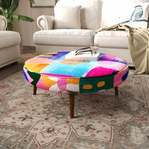 Puede incluir: Un puf redondo con un vibrante patrón abstracto multicolor que presenta tonos de rosa brillante, azul, amarillo y verde. La pieza se apoya sobre cuatro patas de madera cónicas y tiene un libro blanco y un vaso transparente sobre su superficie.