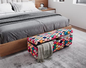 May include: A colorful storage bench with a lid, featuring a pattern of overlapping, multi-colored shapes. The bench is open, revealing folded blankets and a gray scarf. The bench is placed at the foot of a bed with a gray comforter.