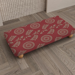 May include: A rectangular footstool with a red fabric top featuring a repeating pattern of beige floral and leaf designs. The footstool is supported by four small, rounded wooden legs. The design is a mix of floral and botanical elements.