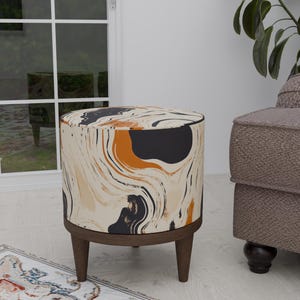 May include: A round, cream-colored ottoman with a black and orange abstract pattern. The ottoman has wooden legs and a dark brown base.