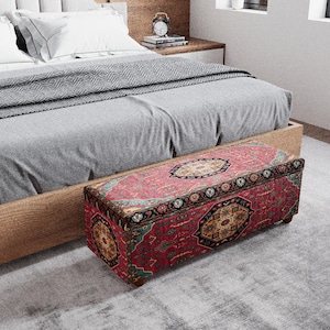 May include: A rectangular ottoman with a red, black, and gold patterned design sits at the foot of a bed. The ottoman has a wooden frame and short legs. The bed has a light wood frame, gray bedding, and white pillows.
