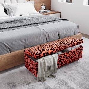 May include: A leopard print storage bench in front of a bed. The bench has an orange background with black spots. A gray blanket and a light gray scarf are visible. The bed has a gray comforter and white pillows.