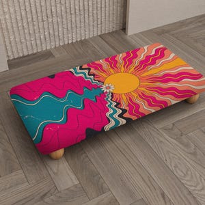 May include: A rectangular footstool with a retro design. The top features a vibrant illustration of a sun with radiating orange and pink rays, and a wavy blue and pink landscape. The stool has four wooden legs.