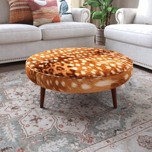 May include: A round ottoman featuring a brown and white spotted faux fur upholstery that mimics the appearance of a deer hide. The piece is supported by four tapered dark brown wooden legs, providing a stable base for this circular furniture item.
