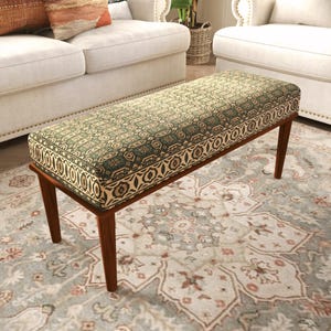 May include: A rectangular upholstered bench featuring a dark wood frame with four tapered legs. The seat is covered in a fabric with a repeating geometric pattern in shades of green, cream, and dark brown, finished with a decorative border.