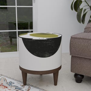 Pode incluir: Um pufe cilíndrico branco com um design abstrato em preto e verde-oliva. O pufe tem uma base e pernas de madeira castanha escura. O design é moderno e minimalista, adequado para a decoração da sala de estar.