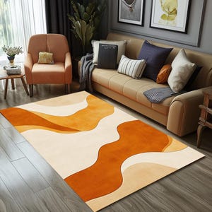 Könnte beinhalten: Ein rechteckiger Teppich mit einem abstrakten Design in Orange-, Creme- und Beigetönen. Der Teppich weist fließende, organische Formen auf, die einen modernen, künstlerischen Look erzeugen. Er liegt auf einem Holzboden in einem Wohnzimmer.