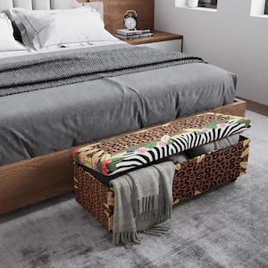 May include: A rectangular storage bench with a zebra, leopard, and floral patterned lid. The bench is open, revealing pillows and a gray blanket. The bench is placed at the foot of a bed with gray bedding.