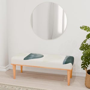 May include: A white bench with a teal and gray abstract design on the seat. The bench has wooden legs and is sitting on a light brown wood floor. A round mirror and a potted plant are in the background.