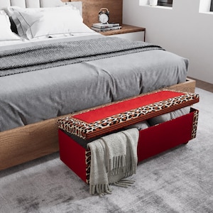 May include: A red storage bench with a leopard print lid is positioned at the foot of a bed with gray bedding. A gray blanket and pillows are visible. The bench is open, revealing folded items.