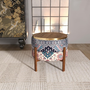 May include: A round ottoman with a dark wood top and a gold metal rim. The base is upholstered in fabric featuring a multi-coloured floral and geometric pattern in shades of blue, pink, and cream. It stands on four straight dark wooden legs.