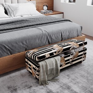 May include: A black and white striped storage bench with a leopard print design. The bench is open, revealing pillows and a gray blanket. The bench is at the foot of a bed with a gray comforter.