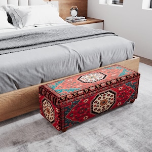 May include: A rectangular storage bench with a red, teal, and cream patterned design. The bench is placed at the foot of a bed with a light wood frame and gray bedding. The bench has small wooden legs and a decorative, patterned top.