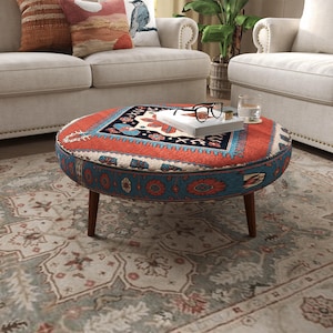 May include: A round ottoman featuring a vibrant orange and blue patterned fabric with intricate geometric and floral motifs. The piece is supported by four dark brown wooden legs and holds a white book topped with black-rimmed eyeglasses.