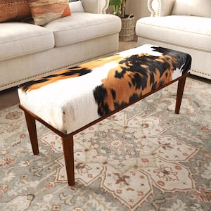 May include: A rectangular bench featuring a cowhide-patterned upholstered seat with a mix of white, black, and brown patches. The bench is supported by four tapered wooden legs in a medium brown finish, providing a stable base for the seat.