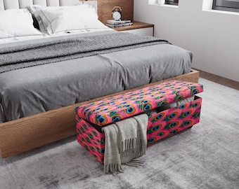 May include: A pink and teal storage bench with a peacock feather pattern sits at the foot of a bed. The bench is open, revealing a gray blanket and other items. A gray throw blanket is draped over the edge.