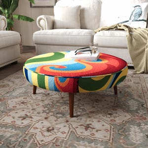 May include: A round ottoman featuring a vibrant, multi-colored fabric upholstery with swirling patterns in red, orange, yellow, green, and blue. The piece is supported by four tapered wooden legs and has a flat top surface.