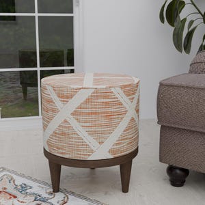 May include: A round, orange and white patterned ottoman with wooden legs. The ottoman is sitting on a white rug with a colorful pattern.