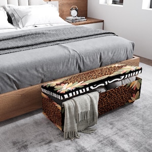 May include: A rectangular storage ottoman with a leopard print design and a black and white floral border. The ottoman is open, revealing folded blankets and pillows. A gray blanket is draped over the edge.