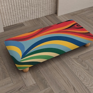 May include: A rectangular footstool with a vibrant, colorful design. The top features a wavy pattern in red, orange, yellow, blue, and green. It has four short, rounded wooden legs. The footstool is placed on a wooden floor.