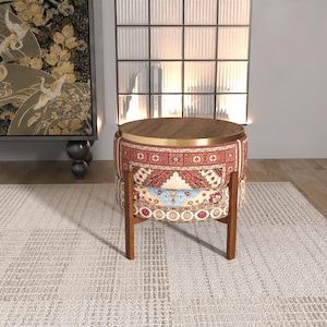 May include: A round ottoman with a dark wood top and a gold metal rim. The base is upholstered in a patterned fabric featuring red, cream, and blue geometric and floral motifs. It is supported by four straight, dark brown wooden legs.