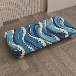 May include: A rectangular footstool with a wave pattern in shades of blue and off-white. The stool has four short, rounded wooden legs. The design evokes a coastal or nautical theme, suitable for home decor.