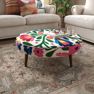 May include: A round ottoman featuring a white fabric upholstery with a vibrant floral pattern in shades of red, pink, blue, and green. The piece is supported by four tapered wooden legs and displays a book and glasses on its flat surface.