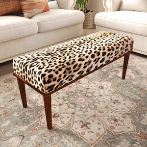 Puede incluir: Un banco rectangular con una parte superior acolchada tapizada en un estampado de leopardo en tonos beige, negro y marrón. El mueble se apoya sobre cuatro patas de madera cónicas con un acabado en marrón medio, ofreciendo una base estable.