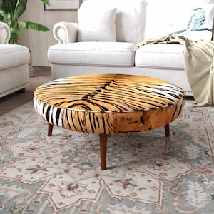 Puede incluir: Un otomano redondo con un patrón de rayas de tigre texturizado en tonos naranja, negro y blanco. La pieza está sostenida por cuatro patas de madera cónicas con un acabado marrón medio, proporcionando una base estable para este mueble bajo.