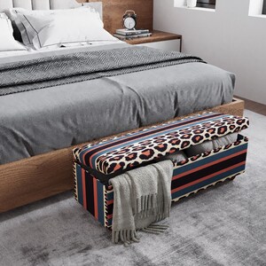 May include: A storage bench with a leopard print top and striped sides sits at the foot of a bed. The bench is open, revealing folded blankets and a gray scarf. The bed has a gray comforter and pillows.