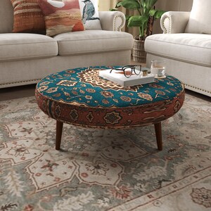 May include: A round ottoman with a teal and rust-colored patterned fabric featuring intricate floral and geometric motifs. The piece stands on four short, tapered wooden legs and supports a white book topped with black-rimmed glasses.