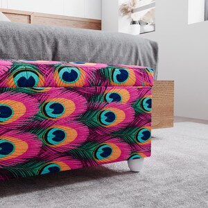 a colorful, patterned ottoman or storage box with a vibrant design of peacock feathers in shades of pink, blue, green, and orange. The ottoman is placed on a gray carpeted floor, and there is a wooden shelf or cabinet visible in the background.