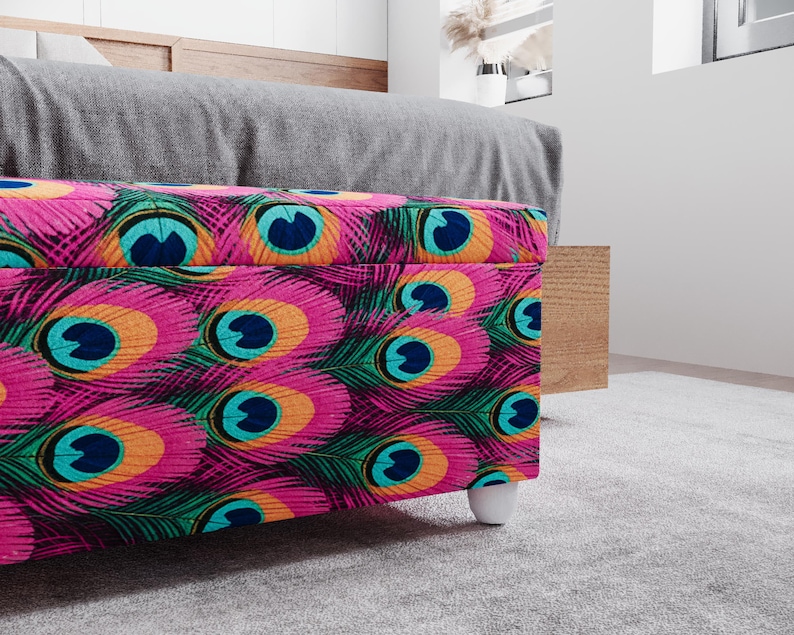 a colorful, patterned ottoman or storage box with a vibrant design of peacock feathers in shades of pink, blue, green, and orange. The ottoman is placed on a gray carpeted floor, and there is a wooden shelf or cabinet visible in the background.