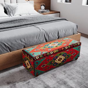 May include: A rectangular storage ottoman with a vibrant, patterned design in red, teal, and gold. The ottoman is placed at the foot of a bed with a gray comforter. The design features geometric shapes and floral motifs.