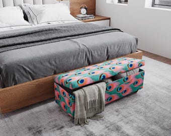 May include: A rectangular storage ottoman with a peacock feather design in teal, pink, and orange sits at the foot of a bed. The bed has a gray comforter and a gray patterned throw blanket. A gray blanket is draped over the ottoman.