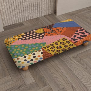 May include: A rectangular footstool with a patchwork design featuring various patterns and colors, including polka dots, floral prints, and animal prints. The stool has wooden legs and a padded top. The color palette includes orange, pink, blue, and yellow.